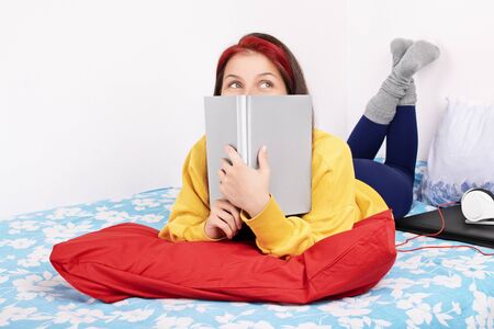 Young beautiful girl lying on the bed in a bedroom, next to a laptop and headphones, holding a book over her face, trying to study but instead she is daydreaming of being somewhere else, isolated on a white background.の写真素材