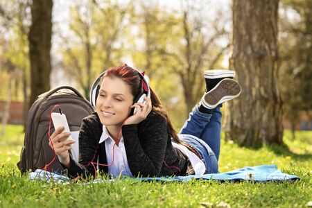Young beautiful girl lying on a blanket on the grass in the park, listening to music and looking at the phone.の写真素材