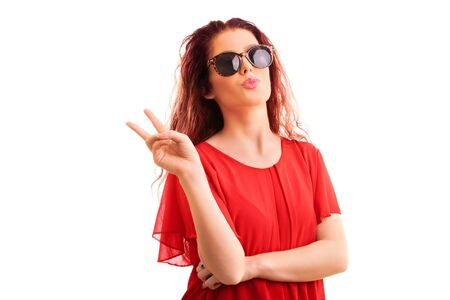 Young beautiful redhead girl in a red blouse with sunglasses gesturing the peace sign and sending a kiss, isolated on a white background.の写真素材