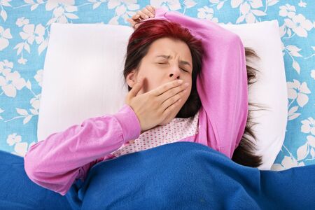 Young beautiful girl lying on a pillow under a blanket yawning, isolated on a white background.の写真素材