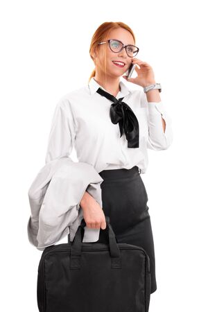 Smiling beautiful young businesswoman with glasses in a suit talking on the phone, holding a laptop bag, isolated on a white background. Business communication concept.の写真素材