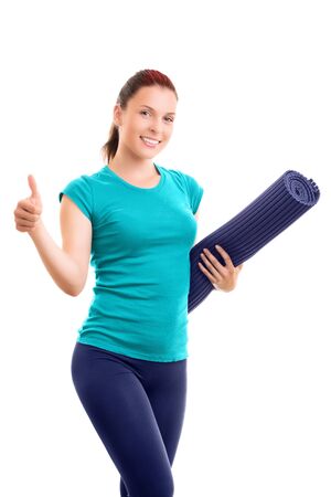 Beautiful smiling young girl in sportswear holding a yoga mat giving thumbs up, isolated on white background. Healthy lifestyle concept. Fitness and yoga concept.の写真素材