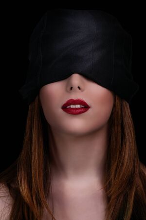 Low key shot of a gorgeous young woman with a black blindfold covering her eyes, isolated on black background. Beautiful blindfolded woman with red lipstick.の写真素材