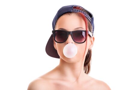 Beautiful stylish girl with backwards snapback cap and sunglasses, blowing a pink bubble with her chewing gum, isolated on white background.の写真素材