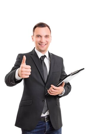 Handsome confident smiling businessman in a suit standing, holding a clipboard and giving thumb up gesture, isolated on white background. Young attractive businessman wearing a suit and tie giving thumb up.の写真素材