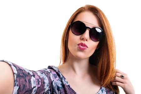 Young beautiful girl with sunglasses taking a selfie while blowing a kiss, looking at the camera POV, isolated on white background. Attractive redhead girl with long hair taking a selfie. Lifestyle, happiness, and social concept.の写真素材