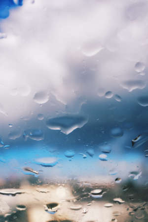 Selective focus of rain drops on a glass window surface with beautiful blurry sky and clouds in the background. Dramatic background concept. Light after the storm.の写真素材