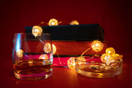 Glass of whiskey and burning cigarette resting on an ashtray, with stack of books and lights in the background. Late night indulgences. Writersâ bad habits. Alcohol and smoking concept.の写真素材