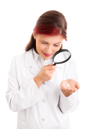 Beautiful smiling young doctor in white lab coat examining a white pill though a magnifying glass, isolated on white background. Scientist with magnifying glass examining pill. Medication scrutiny concept.の写真素材
