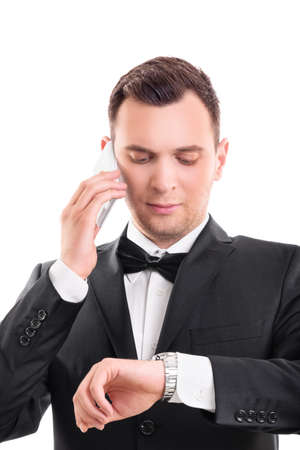 Close up portrait of a handsome serious man in black suit with bow tie talking on mobile phone while looking at his wristwatch, isolated on white background. Late concept.の写真素材