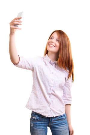 Portrait of a beautiful smiling young woman in smart casual clothes taking a selfie with her mobile phone, isolated on white background. Social media, communication concept.の写真素材