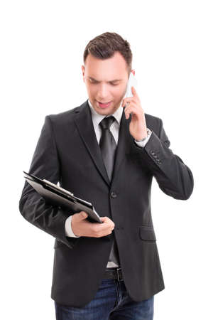Portrait of a young fashionable businessman, looking at a clipboard and talking on a mobile phone, looking worried, pensive, stressed, serious, anxious, isolated on white background.の写真素材