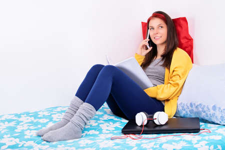 Beautiful young girl sitting on her bed in her room holding a book and talking on a mobile phone. Young girl in bed smiling and talking on the phone. Communication, friendship, quarantine concept.の写真素材