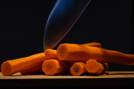 Carrots laying on a wooden cutting board, with a black background. A knife seems to start cutting the vegetables. Low-key photograph.の写真素材
