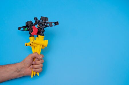 The male hand holds a yellow plastic supports rack and a sprinklers on a blue background. Gardening...
