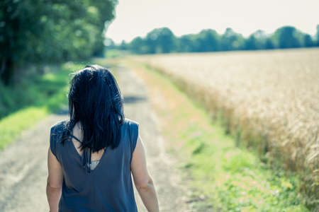 Young woman with black hair is walking along a dirt road between forest and wheat field in sunny...