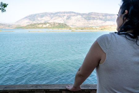 Young woman with black curly hair and gray dress looks at the turquoise colored sea with mountainsの写真素材