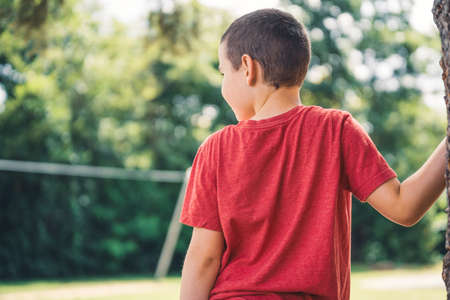 Boy in a red shirt leans against a tree in the playground and looks at the cable carの写真素材