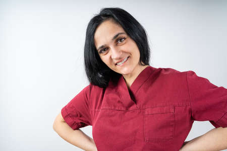 Black haired young nurse in burgundy colored hospital clothing looks smiling at the cameraの写真素材