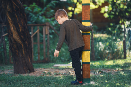 Cute little boy in a sweatshirt, sweatpants and slippers walks around a colorful wooden post in the gardenの写真素材