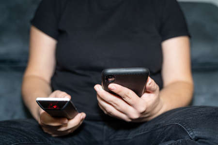Young woman sits on the couch and holds the remote control in one hand and the mobile phone in the...