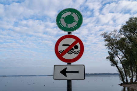 Green sign with a swimming ring, red sign with a crossed out swimmer and white sign with an arrow pointing to the right on a post at the lake beachの写真素材
