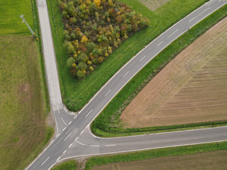 Shot from above of a road crossing between trees, grass and plowed agricultural fields in the autumn...