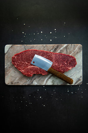 Raw beef steak with meat cleaver on dark background, top view