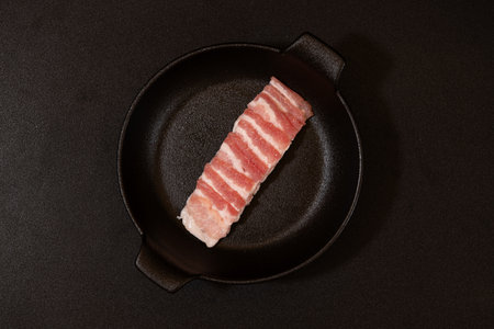 Raw bacon on a black frying pan, isolated on black background.