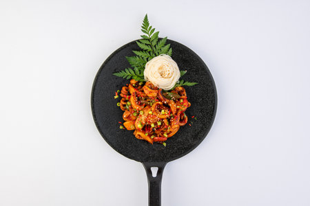 Spicy stir-fried noodles with vegetables and sesame seeds in black frying pan on white background.