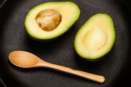 Halved avocado on a black plate with a wooden spoon