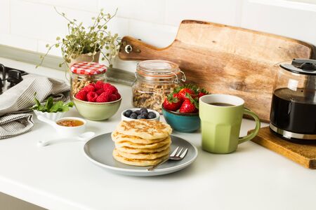 Pancakes with berries and honey on a gray plate and with a cup of coffee on a kitchen counter