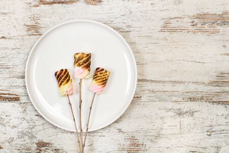 Yellow and pink burned marshmalows on sticks on a plate and on a wooden tableの写真素材