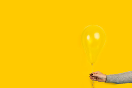 Woman hand with black nails holding a yellow balloon on a yellow backgroundの写真素材