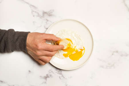 Man hand with a slice of bread and a broken fried egg on a white plateの写真素材