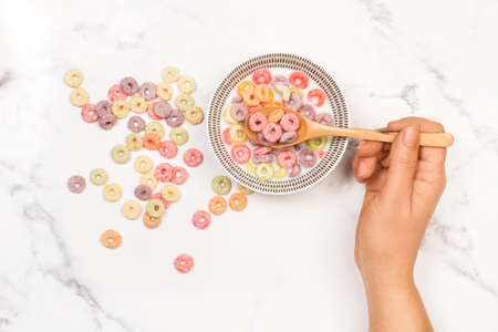 Woman hand holding a wooden spoon on bowl with fruit flavor colored cereals with milk on a kitchen marble counterの写真素材
