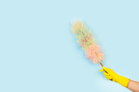 Woman hand with yellow rubber glove holding a multicolor duster on a light blue background with copy...