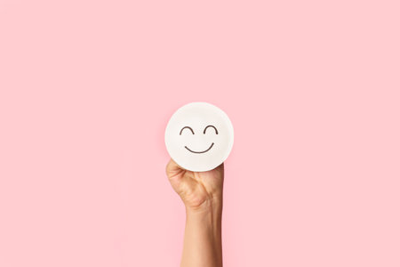 Woman's hand holding a round paper in the shape of a happy face on a pink backgroundの写真素材