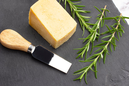 A piece of parmesan cheese, a knife and rosemary twigs on a black slab in a top view