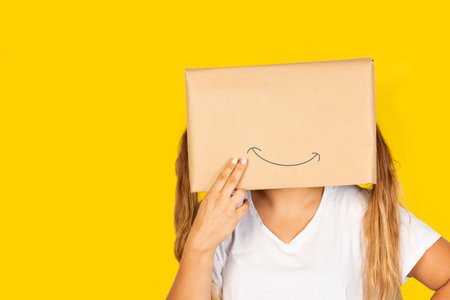 Woman with her head in a box and happiness gesture drawn on it and standing on a yellow background with copy spaceの写真素材