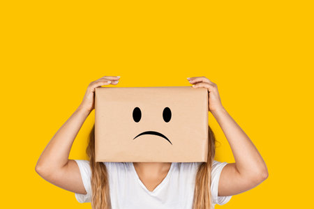 Woman with her head in a box, hands on it and sadness gesture on a yellow background with copy spaceの写真素材