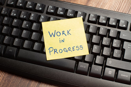 A black computer keyboard and a yellow paper note with work in progress phrase on a wooden desk