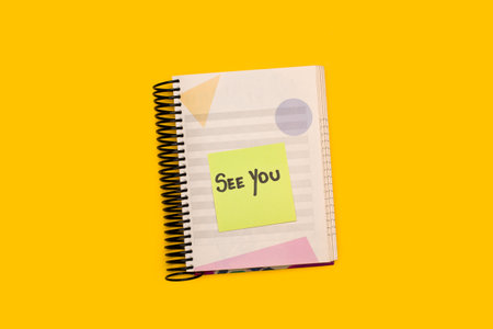 A spiral notepad with a sticky paper with See You short phrase handwritten on it on a yellow backgroundの写真素材
