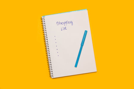 Shopping list handwritten on a white page of a notepad and a blue marker on a yellow background with copy spaceの写真素材