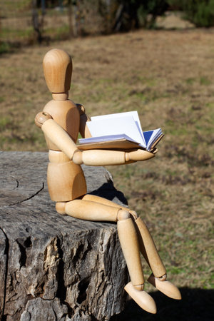 A wooden mannequin reading a book on his hands sitting on a tree trunk in a gardenの写真素材