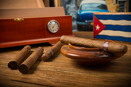 Cuban cigars and humidor with ashtray on rustic wooden table with Cuban painting of american old car in backgroundの写真素材