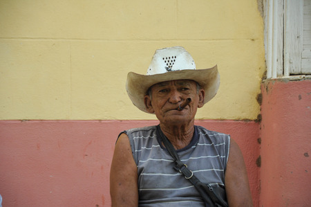 TRINIDAD, CUBA - JANUARY 28 , 2013 Cuban local man smoking cigar and posing to photos while sitting on street in UNESCO protected city of Trinidad, Cuba.のeditorial素材