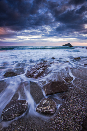 Red rocks on beach at sunset, dramatic clouds and wavesの写真素材