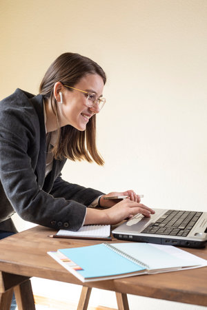 A smiling young woman with glasses works with her laptop. She wears headphones and listens to a video call.の写真素材