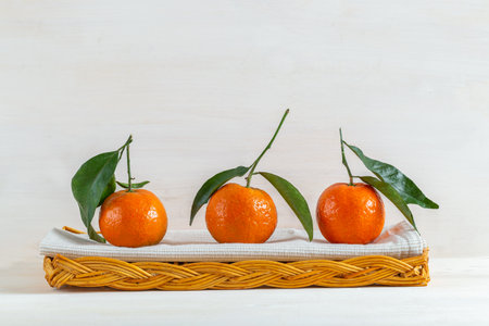 A still life of three fresh tangerines, front view, side by side, in a wicker basket with a kitchen towel. Tangerines have their stem and their leaves. The background is white.の写真素材
